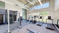 Located at 10000 West Missouri Avenue in Glendale, Arizona, this modern fitness center is designed to support a comprehensive workout experience in a well-appointed, single-room facility. The space is defined by its striking double-height ceilings with exposed structural elements and visible ductwork, lending an industrial-contemporary aesthetic that feels open and energizing.

Floor-to-ceiling windows line multiple walls, flooding the interior with abundant natural light and offering views of the surrounding landscaped grounds and mature trees. The neutral-toned carpet flooring provides comfortable underfoot support throughout the workout area.

The fitness center is equipped with a well-rounded selection of equipment. Cardio offerings include treadmills, elliptical machines, and stationary spin bikes, several of which feature integrated digital displays. A multi-functional cable and rack system anchors the strength training area, complemented by a tiered dumbbell rack, adjustable weight benches, and functional training tools such as a heavy tire and plyo box. A full-length mirror along one wall enhances the sense of space and supports form-check during training.

Bold, geometric wall murals in teal, navy, and white add a modern artistic touch, while ceiling fans and a well-planned lighting system ensure a comfortable environment. The space presents a clean, well-organized layout suitable for a wide range of fitness activities.