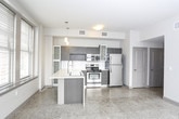 This contemporary single-floor residence offers a well-designed layout that balances modern aesthetics with functional living. The open-concept kitchen serves as the centerpiece of the main living area, showcasing dark gray cabinetry with frosted glass upper panels, a full suite of stainless steel appliances including a gas range, built-in microwave, and refrigerator, and a striking white quartz waterfall island with an integrated sink. Polished terrazzo flooring throughout the kitchen and main area adds a sophisticated, industrial-modern character while reflecting abundant natural light from the large, street-facing windows fitted with horizontal blinds. The bedrooms feature plush wall-to-wall carpeting and generous windows offering urban skyline views, providing a calm and comfortable retreat. One bedroom includes a walk-in closet alcove, adding practical storage. The bathroom is finished with polished terrazzo flooring, a white vanity with under-cabinet storage, a framed mirror, and a tub-shower combination set against tile surround, all accented by bold sage-green walls that lend a distinctive, energetic character. The overall ambiance of the unit is clean, modern, and urban, with thoughtful material choices throughout.