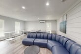 Located at 333 Holly Creek Ct in Spring, TX, this single-floor community recreation room offers a well-designed, multipurpose living space built for both relaxation and entertainment. The room features light gray walls, wood-look plank flooring, and recessed lighting throughout, creating a bright and welcoming atmosphere.

At the heart of the space sits a bold, curved blue sectional sofa that serves as the primary lounge focal point, complemented by a round coffee table and a neutral accent chair. A wall-mounted flat-screen TV is positioned above a slim console table, creating a comfortable viewing area. The room also accommodates two full-size ping-pong tables, making it an ideal venue for recreational activities and social gatherings.

A shiplap accent wall adds a modern yet cozy architectural detail, while abstract wall art in cool blue tones enhances the visual appeal. Multiple windows dressed with white blinds allow natural light to filter in. Additional seating includes built-in bench seating along one wall and two small café-style tables with chairs, providing flexible options for dining or casual conversation. A wall-mounted mini-split HVAC unit ensures year-round comfort. The overall design balances function and style, making this a versatile and inviting community gathering space.