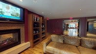 This single-floor lounge is a well-appointed communal space designed to accommodate both relaxation and collaborative work. The interior is defined by rich burgundy accent walls contrasted against a crisp white ceiling with recessed lighting, creating a warm yet contemporary ambiance throughout. Warm-toned hardwood flooring runs continuously across the entire space, reinforcing a cohesive and inviting atmosphere.

The seating area serves as the social focal point, anchored by a built-in fireplace with a tile surround and a wall-mounted flat-screen television above it. Flanking the fireplace are tall wooden built-in shelving units displaying decorative objects, adding character and depth to the wall. A generously sized sectional sofa and additional seating are arranged around a dark coffee table, creating an ideal setting for casual gatherings.

Adjacent to the lounge area, a long communal work table with industrial-style seating provides a functional workspace, complemented by additional built-in shelving along the opposite wall. Natural light pours into the space through a series of large glass French doors that open directly onto an outdoor patio, offering views of lush greenery and creating a seamless indoor-outdoor connection. Window shutters offer privacy when desired.
