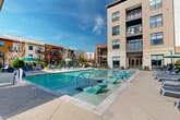 Located at 1308 Lorna St in Charlotte's vibrant 28205 corridor, this outdoor amenity space is centered around a stunning resort-style swimming pool that serves as the focal point of the community. The pool features distinctive in-water lounge chairs finished in aqua teal, allowing residents to relax partially submerged while enjoying the open sky above. The surrounding deck is generously proportioned, offering abundant space for sunbathing with rows of blue chaise lounges and colorful umbrellas providing shade throughout the day.

A thoughtfully designed covered cabana structure anchors one side of the pool area, featuring ceiling fans, an outdoor fireplace, built-in grilling stations, and a wet bar — creating an ideal space for outdoor entertaining in all weather conditions. The pergola-style roof allows airflow while providing ample shade, and the warm wood-panel siding adds a natural, contemporary aesthetic.

Mature Italian cypress trees and manicured hedgerows line the perimeter, providing privacy and a lush green backdrop. The multi-story residential building rises directly adjacent to the pool, with private balconies overlooking the amenity area. The overall ambiance is polished and resort-inspired, combining modern architecture with thoughtfully curated outdoor living elements in the heart of Charlotte.