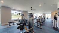 This expansive fitness center is thoughtfully designed to accommodate a full range of workout styles across distinct functional zones. The space is organized to separate cardio equipment, strength machines, and free-weight areas, allowing users to move efficiently through their routines. Large floor-to-ceiling windows line the perimeter, flooding the interior with natural light and offering open views of the surrounding landscape, creating an inviting and energizing atmosphere throughout the day.

The cardio zone is equipped with a row of treadmills and Spinning bikes positioned directly in front of the oversized windows, maximizing the outdoor views during workouts. Adjacent to this area, a comprehensive selection of Precor selectorized strength machines is arranged to provide targeted muscle training. A wall-mounted flat-screen television adds entertainment options in the strength training zone.

The free-weight area features a full dumbbell rack, adjustable benches, stability balls, and floor space suitable for stretching or functional training. Full-length mirrors reflect the room and enhance the sense of openness. Recessed lighting and ceiling fans are installed throughout, ensuring consistent illumination and airflow. The flooring combines dark carpet tiles with lighter textured panels, clearly delineating each workout zone. Glass-paneled entry doors connect the fitness center to adjacent amenity spaces.