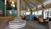 This single-story space makes a strong architectural impression through its soaring vaulted ceiling with exposed wood beam detailing, creating an airy, open atmosphere throughout the interior. A central structural column anchors the space, surrounded by a distinctive circular bench upholstered in bold horizontal stripes of brown, tan, and cream — serving as both a functional seating element and a visual focal point.

The open layout is divided into distinct functional zones. A lounge or waiting area near the entrance features comfortable armchairs arranged on patterned carpeting, while a counter-height reception or service area lines the far wall, finished in a teal-blue tone that adds a splash of color against the neutral surroundings. To the right, a workstation or business area offers seating with task lighting provided by classic lamp fixtures.

Natural light filters through multiple windows along the perimeter, complementing a cluster of pendant lights that hang from the vaulted ceiling, adding warmth to the space. The overall ambiance is contemporary yet inviting, blending thoughtful design details with practical functionality suited to a communal or hospitality-oriented environment.