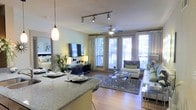This single-floor residence showcases an open-concept layout that connects the kitchen, dining, and living areas into one cohesive, functional space. The kitchen features granite countertops with a breakfast bar overhang, accommodating casual dining with place settings already in view. A stylish pendant light hangs above the bar, complementing the track lighting overhead that provides targeted illumination throughout the space.

The living area is anchored by a plush sectional sofa, a glass-top coffee table, and a soft area rug layered over warm hardwood flooring. A ceiling fan with a light fixture is centrally positioned above, promoting airflow and comfort. The entertainment setup includes a wall-mounted flat-screen TV atop a sleek media console, flanked by decorative greenery that adds a natural touch.

Large floor-to-ceiling windows and glass doors line the far wall, flooding the interior with natural light and offering views of outdoor greenery. Sheer curtains frame the windows elegantly. The overall ambiance is contemporary and inviting, combining neutral tones, modern furnishings, and thoughtful lighting to create a comfortable and well-appointed living environment.
