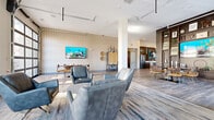 Located at 644 Chlois Ln in Columbus, Ohio, this single-floor amenity space offers a thoughtfully designed environment that blends modern aesthetics with functional versatility. The open-concept layout is unified by wood-look luxury vinyl plank flooring that flows seamlessly throughout, complementing a contemporary color palette of warm neutrals, deep walnut tones, and vibrant teal accents.

The main lounge area features plush leather seating arranged around a central gathering space, with a geometric-patterned accent wall serving as a backdrop for a wall-mounted flat-screen television. An adjacent built-in shelving unit displays curated décor alongside an additional television, creating an inviting focal point. A separate seating nook with floor-to-ceiling windows offers city views and bar-height workspace seating alongside a built-in daybed, ideal for quiet work or relaxation.

The kitchen area is outfitted with stainless steel appliances, a large stone-topped island with textured panel detailing and bar seating, dark cabinetry with open shelving accented in teal, and a full-size refrigerator. A private meeting room features a glass sliding barn door, a fireplace, and direct access to an outdoor area. Track lighting and recessed fixtures ensure the entire space is well-lit throughout.