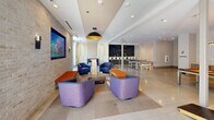 Located at 125 Anderson Hwy in Clemson, South Carolina, this single-floor amenity space offers a well-appointed community gathering area designed for both relaxation and recreation. The space is divided into two distinct zones that flow seamlessly into one another.

The lounge area showcases a refined aesthetic with a textured stone accent wall serving as a striking backdrop for a large wall-mounted flat-screen television. Comfortable seating — including bold blue and purple upholstered armchairs paired with orange bench ottomans — is arranged around a rustic-modern coffee table, creating an inviting social atmosphere. A large pendant light with a black and gold dome finish anchors the space, while recessed lighting and wall sconces provide warm, layered illumination. Natural light filters in through windows, adding brightness to the room.

Transitioning into the game room, polished concrete flooring replaces the carpeted lounge area, reflecting the space's more active purpose. Amenities include ping pong tables, a cornhole board, and a pool table, all set beneath a dramatic chandelier and surrounded by crisp white walls accented with a bold chalkboard feature wall. A bar-height counter with seating lines one side, offering a casual spot for spectators. Together, the two areas create a dynamic, energetic environment ideal for community engagement and social interaction.