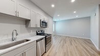 This single-floor unit offers a clean, contemporary design with a cohesive neutral color palette and luxury vinyl plank flooring that runs throughout the entire space. The kitchen is well-appointed with soft gray shaker-style cabinetry, light stone-look countertops, a stainless steel sink, and a full suite of stainless steel appliances including a refrigerator, electric range, over-the-range microwave, and dishwasher. Recessed lighting illuminates the kitchen and flows into the adjacent open living area, which provides generous room for flexible furniture arrangements. Natural light enters through a window, enhancing the bright and airy atmosphere. The bedroom features wood-tone vinyl plank flooring, a dedicated closet with wire shelving, and an electrical panel conveniently integrated into the room. An additional room offers a versatile, open layout suitable for various uses. The bathroom is finished with clean white tile surrounds, a built-in niche shelf in the tub/shower combo, and a minimalist design. A solid entry door with a keypad lock provides secure access, and the space is equipped with a fire safety alarm system, underscoring its attention to modern safety and convenience standards.
