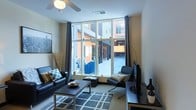Situated in the heart of historic Savannah, Georgia, this single-floor residence at 646 W Bay Street blends contemporary design with urban convenience. The living area serves as the centerpiece of the space, featuring large, multi-pane floor-to-ceiling windows that flood the room with natural light and offer views of the surrounding cityscape. The windows are framed by elegant dark curtains, adding a refined contrast to the bright, neutral walls. A ceiling fan overhead provides both function and a casual architectural detail. The living room is thoughtfully appointed with modern furnishings, including a sleek black leather sofa and a coordinating accent chair, arranged around a geometric-patterned area rug. A low-profile coffee table and media console complete the entertainment setup, creating a cohesive and comfortable gathering space. The overall ambiance is clean, modern, and inviting, with a palette of neutral tones accented by contemporary décor. The open layout maximizes the sense of space, making this Savannah residence an ideal urban retreat that balances style and everyday functionality.