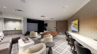 This sophisticated golf simulator lounge offers a well-appointed, single-level entertainment space designed to blend sport and leisure seamlessly. The room is anchored by a full indoor golf simulator bay, complete with a projection system, hitting mat, and a large screen that serves as the focal point of the space. Recessed lighting throughout the smooth white ceiling ensures consistent, even illumination across the entire area.

The lounge is furnished with multiple distinct seating configurations, including a cognac leather sofa, plush bouclé curved armchairs, and a taupe sectional, all arranged atop a bold plaid-patterned area rug that adds a classic, clubhouse-inspired character to the interior. Sculptural white coffee tables and dark side tables complement the eclectic yet cohesive furniture selection.

Along one wall, a bar-height counter with dark wicker stools provides additional seating beneath dual wall-mounted flat-screen TVs, ideal for viewing and socializing. Textured wall panels in warm taupe tones line key sections of the room, lending depth and warmth. A decorative starburst wall accent and a small wet bar with cabinetry further enhance the upscale, club-like ambiance. The overall aesthetic is modern and refined, thoughtfully designed for both active recreation and relaxed social gatherings.