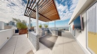 Located at 915 West Grape Street in San Diego's vibrant Little Italy neighborhood, this rooftop patio is a thoughtfully designed outdoor retreat that delivers an exceptional urban living experience. The space is defined by its commanding panoramic views of San Diego Bay and the downtown skyline, visible from nearly every corner of the terrace.

The rooftop is organized into distinct functional zones. A fully equipped outdoor kitchen anchors one section of the patio, constructed with gray brick detailing and topped with sleek countertops. It features stainless steel appliances including a built-in grill, refrigeration, and ample prep space, all sheltered beneath a handsome wood-slatted pergola that adds warmth and architectural character.

Adjacent to the kitchen, a generously sized lounge area is outfitted with contemporary outdoor seating, including low-profile sofas with navy and orange cushions arranged around built-in concrete fire pit tables — ideal for evening gatherings. Oversized planters in weathered Corten steel house mature olive trees and ornamental plantings, providing natural greenery and subtle privacy screening.

A separate decked zone with light wood flooring features casual bean bag seating and additional lounge chairs, creating a relaxed, resort-like atmosphere. The perimeter is framed by horizontal metal railings, allowing unobstructed sightlines to the bay, skyline, and surrounding cityscape. Bold circular wall art adds a playful, curated touch to the space's modern aesthetic.