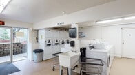 Located at 8684 SW Borders St in Portland, Oregon, this single-floor shared amenity facility serves as a centralized laundry and mail center. The space is bright and functional, illuminated by fluorescent overhead lighting throughout, with light-toned walls and durable speckled flooring that contribute to a clean, utilitarian aesthetic.

The laundry area is well-equipped with a generous bank of top-loading washing machines and a matching row of stacked dryers, all coin-operated and prominently featured along the walls. A freestanding utility sink and rolling laundry carts are also available for resident use, adding to the convenience of the space. Paper towel dispensers are mounted on the walls nearby. The area is designated as a non-smoking zone, clearly marked with signage.

The mailroom section features an extensive array of individual resident mailboxes lining two walls, providing ample storage for incoming mail. A fire extinguisher and fire safety signage are visibly placed for safety compliance.

An additional common area connects the two zones, housing a snack vending machine, a beverage vending machine, and a ceiling fan, creating a small break-like space. A glass entry door allows natural light into the room and provides direct access to the exterior. Overall, the facility is practical and well-organized, designed to meet the everyday needs of its residents efficiently.