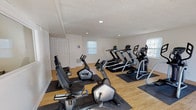 Located at 3550 Pimlico Pkwy in Lexington, Kentucky, this fitness center is a well-equipped, single-floor facility designed to support a well-rounded workout routine. The main exercise area features light wood flooring and a textured ceiling with recessed lighting, creating a bright and clean environment. Multiple windows allow natural light to fill the space, contributing to an open and inviting atmosphere. The cardio zone is outfitted with a generous selection of machines, including treadmills, elliptical trainers, a stair climber, and both upright and recumbent stationary bikes, all positioned on protective floor mats. A glass entry door provides direct access from the exterior, adding convenience and additional light to the main floor. A separate adjoining room serves as a dedicated strength training area, featuring a full rack of dumbbells in varying weights alongside a workout bench, making it suitable for a range of resistance exercises. This room includes a large wall-mounted mirror, enhancing the sense of space and allowing users to monitor their form. The overall layout is functional and efficient, with clearly defined zones for cardio and strength training that cater to a variety of fitness needs.