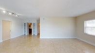 This single-floor space presents a clean, open layout designed for functional everyday living. The main living area is finished with large-format, light-toned ceramic tile flooring that extends throughout, creating a cohesive and easy-to-maintain surface underfoot. Neutral beige walls and white baseboards contribute to a bright, understated aesthetic that suits a variety of furnishing styles.

Ceiling-mounted HVAC vents are positioned throughout, ensuring consistent climate control across the space. The smooth, white ceiling adds to the overall sense of openness in the room.

Toward the rear of the living area, a partial wall division leads into additional spaces. A glimpse of the kitchen is visible, featuring dark cabinetry that provides a warm contrast to the otherwise light interior. Bi-fold closet doors are also visible along the back wall, suggesting convenient built-in storage options nearby.

A doorway connects the living area to what appears to be a hallway, providing access to the remaining rooms of the unit. Overall, the space offers a straightforward, well-maintained environment with a neutral palette that serves as a versatile backdrop for personalization.