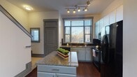 Located at 201 Adair Dr in Chapel Hill, NC, this two-floor residence offers a thoughtfully designed layout with a blend of practical finishes and comfortable living spaces. The kitchen serves as a standout feature, equipped with granite countertop surfaces, a central island, white cabinetry extending to the ceiling, a dark tile backsplash, and black appliances including a range, built-in microwave, and refrigerator. A large window above the sink invites natural light and outdoor views, while track lighting overhead ensures excellent illumination throughout. Rich, dark hardwood flooring runs through the main living areas on the lower level, adding warmth and visual continuity. The open living area is generously proportioned, with clean white walls, gray accent baseboards, and ample electrical outlets along the perimeter. The bathroom features a vessel sink set in a granite vanity, tile flooring, and a glass-enclosed shower with tasteful dark tile surround. Upper-level rooms are finished with plush carpeting, ceiling fans, and large windows that frame views of the surrounding landscape. Bold gray crown molding provides a clean, defined border throughout the upper floor. The consistent use of gray trim, granite surfaces, and hardwood flooring creates a cohesive aesthetic across both levels.