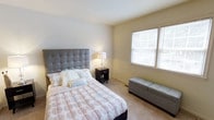 This single-floor space includes a well-appointed bedroom that combines functionality with a clean, understated aesthetic. The bedroom features soft, wall-to-wall carpeting that adds warmth underfoot, complemented by neutral-toned walls that create a calm and restful atmosphere. A prominent double window fitted with white blinds allows natural light to filter into the room, enhancing its open and airy feel. The room comfortably accommodates a queen-sized bed with a tufted upholstered headboard, flanked by a dark-finished nightstand that provides convenient bedside storage. A storage bench at the foot of the bed offers additional functionality. Recessed lighting in the ceiling provides ambient illumination, supplemented by a bedside table lamp for task lighting. Adjacent to the bedroom entrance, a louvered bi-fold closet door provides accessible and well-organized storage space. The hallway leading to the bedroom features crisp white trim and clean wall finishes, maintaining a consistent and polished look throughout. Wall art adds a subtle decorative touch, contributing to the overall cohesive and tidy presentation of the space.