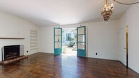This single-floor space exudes a distinctive Spanish Colonial character, blending classic architectural details with warm, inviting interiors. Rich, dark hardwood floors run throughout, anchoring each room with a sense of depth and tradition. The main living area is a generously proportioned room, highlighted by striking teal-framed French doors that open directly onto a lush, plant-filled courtyard. A crystal chandelier adorns the ceiling, adding an elegant focal point, while built-in white shelving lines one wall, offering functional display space. Natural light streams in through the French doors, illuminating the open interior. A hallway with a dramatic barrel-vaulted ceiling connects the living areas, lending an airy, cathedral-like quality to the passage. This hall leads to additional rooms, including a bathroom that continues the Spanish aesthetic with terracotta tile floors, a classic soaking tub, white subway tile walls accented with decorative patterned inserts, and its own barrel-vaulted ceiling. The kitchen features white cabinetry, open shelving, wood countertops, and teal tile detailing around the sink that echoes the color palette found throughout. The private courtyard entrance, framed by climbing greenery and terracotta tile, completes the cohesive, character-rich design of this thoughtfully detailed space.