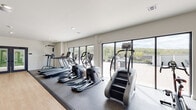 This contemporary fitness center is thoughtfully designed to support a full range of workout routines in a bright, open environment. The space is organized into distinct functional zones, seamlessly connected by a combination of durable rubber flooring and smooth hardwood-style surfaces. The cardio area is generously appointed with multiple treadmills, elliptical trainers, and a stair climber, all positioned to take full advantage of the expansive floor-to-ceiling windows that line one wall. These large-format windows flood the space with natural light and frame sweeping views of a lush, tree-lined landscape and an adjacent outdoor terrace, creating an energizing atmosphere during workouts. The strength training zone occupies a separate but connected area, outfitted with a cable crossover machine, a power rack loaded with weight plates, a flat bench, and several selectorized resistance machines suited for targeted muscle training. A stability ball and additional free weight options round out the functional fitness offerings. The interior features clean white walls, recessed lighting, and a neutral color palette that keeps the aesthetic modern and uncluttered. Double glass-panel doors provide access between zones, contributing to an open, airy flow throughout the facility.