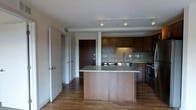 This single-floor space centers around an open-concept kitchen designed for both functionality and style. The kitchen is anchored by a substantial island with a granite countertop, providing ample workspace and serving as a natural divider between the kitchen and surrounding living areas. Dark espresso-toned cabinetry lines the walls, offering generous storage, while a decorative mosaic tile backsplash in warm earth tones adds texture and visual interest between the upper and lower cabinets. Stainless steel appliances, including a range and sink, complete the kitchen's modern, cohesive aesthetic. Warm hardwood flooring extends throughout the space, lending continuity and warmth. Track lighting mounted on the ceiling provides directed illumination across the kitchen area. A hallway visible from the kitchen leads to additional rooms, with multiple doors suggesting separate private spaces beyond. The overall layout feels open and practical, with clean lines and a neutral color palette that creates a welcoming, move-in-ready environment suited to everyday living.