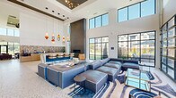 Located at 5705 Apex Dr in Fishers, Indiana, this contemporary community clubhouse delivers a sophisticated, amenity-rich environment designed for both socializing and relaxation. The single-floor layout encompasses a generously sized living area that benefits from dramatic double-height ceilings, which lend an airy, open quality throughout the space.

Floor-to-ceiling windows with bold black steel framing line multiple walls, flooding the interior with abundant natural light and providing sweeping views of the surrounding landscape and community. Industrial-style glass partition walls further enhance the sense of openness, visually connecting distinct lounge zones without sacrificing definition.

The main living area is thoughtfully appointed with multiple seating arrangements, including large modular sofas, accent chairs in leather and upholstered finishes, and glass-topped coffee tables with gold metallic accents. A statement feature wall clad in textured tile panels serves as an anchor for a mounted flat-screen display, flanked by tall potted tropical plants. Polished terrazzo flooring and large-format tile run throughout, adding a refined, modern aesthetic.

A dedicated recreation area housing a billiards table adds a leisure-focused element to the space. Tall open shelving units display curated décor, while a stylish pendant chandelier punctuates one zone. The overall ambiance is clean, modern, and welcoming—ideal for community gatherings.