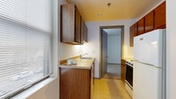 This single-floor unit offers a straightforward, functional layout designed for comfortable everyday living. The living area is bright and open, featuring neutral carpet flooring, white walls, and a baseboard heating system running along the perimeter. Multiple windows dressed with horizontal blinds allow natural light to filter in, creating an airy atmosphere throughout the space.

The galley-style kitchen is efficiently laid out with warm-toned wooden cabinetry lining both walls, providing ample storage above and below. It is equipped with a stainless range, a refrigerator, and a single-basin sink set into a countertop. The light vinyl tile flooring contrasts neatly with the cabinetry and connects seamlessly to the living area through an interior doorway.

The bedroom is generously proportioned with soft carpet underfoot, white walls, and a closet with classic panel doors. A window with blinds brings in natural light, and a baseboard heater ensures year-round comfort. A separate entry door leads directly into this room, offering added flexibility.

The bathroom is clean and well-appointed, featuring light tile flooring, a single-sink vanity with cabinet storage, a toilet, and a walk-in shower enclosed with a frosted glass door and chrome frame. The overall design of the unit is practical and move-in ready.