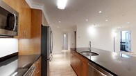 This single-floor, 2-bedroom, 2-bathroom residence offers a thoughtfully designed layout with modern finishes and a cohesive aesthetic throughout. The kitchen is a standout feature, showcasing warm medium-tone wood cabinetry that provides ample storage, complemented by sleek dark granite countertops. A central island with an integrated stainless steel sink adds both functionality and a visual focal point. Stainless steel appliances, including a full-size refrigerator, complete the contemporary kitchen ensemble. Recessed lighting and a flush ceiling fixture ensure the space is well-lit, while crown molding along the ceiling adds a refined architectural detail. Light-toned hardwood flooring flows seamlessly from the kitchen through the hallway, lending warmth and continuity to the interior. The corridor connects the main living areas to the two bedrooms, maintaining an efficient and open flow. White walls throughout keep the atmosphere bright and neutral, offering a versatile backdrop for any furnishing style. The overall ambiance is clean, modern, and move-in ready, making this residence a practical and stylish living space.