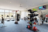 Located at 1111 Oakley Industrial Blvd in Fairburn, GA, this single-floor fitness center offers a spacious and thoughtfully designed workout environment. The open layout is finished with durable blue tile-pattern carpeting underfoot and features smooth, neutral-toned walls accented by recessed lighting and ceiling fans that maintain a bright, comfortable atmosphere throughout.

The facility is comprehensively equipped to support a wide range of fitness activities. A dedicated cardio zone along one wall houses treadmills and elliptical machines, while a large wall-mounted mirror and mounted flat-screen TV add both functionality and entertainment. The free weights section includes an extensive dumbbell rack, kettlebells, and medicine balls arranged on a multi-tier stand for easy access. Functional training is well-represented with a heavy bag boxing station, TRX suspension system, and a cable machine.

Strength training is supported by a multi-use weight machine and adjustable benches. Spinning bikes and aerobic step platforms occupy a separate area near the entrance corridor, making the space versatile for group or individual training. High-set windows along the perimeter walls allow natural light to filter in, creating an open and energizing environment. Wall-to-wall mirrors further enhance the sense of space and allow users to monitor their form throughout their workout.