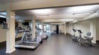 This fitness center offers a well-organized and spacious workout environment designed to accommodate a variety of exercise routines. The room features durable rubber flooring throughout, providing a practical and safe surface for all types of physical activity. Overhead fluorescent lighting ensures the space is consistently well-lit, while large windows at the far end of the room allow natural light to filter in, creating a bright and inviting atmosphere.

The cardio section is prominently featured along one side of the room, housing multiple treadmills, elliptical trainers, and a stair-stepper machine. A wall-mounted television is positioned nearby, offering entertainment during workouts. Structural columns are integrated throughout the space and complement the clean, neutral color palette of the walls and ceiling.

Toward the rear of the room, a cable and weight machine provides options for strength and resistance training, giving users a comprehensive workout experience. The open layout ensures ample circulation space between equipment, reducing congestion during peak usage. Overall, the fitness center presents a functional, no-frills environment suited for both cardio and strength-focused training.
