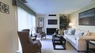 This single-floor space is thoughtfully designed with a clean, contemporary aesthetic that balances comfort and style. The living area serves as the centerpiece, showcasing a classic fireplace framed by a sleek wall-mounted television above it, creating a natural focal point for the room. Large windows dressed with elegant green valances and sheer white curtains allow an abundance of natural light to fill the space, enhancing its bright and airy atmosphere. The interior features a neutral color palette of warm beiges and whites, accented by rich brown leather wingback chairs and a crisp white sofa adorned with light blue decorative pillows. A dark espresso coffee table with a lower shelf anchors the seating arrangement, offering both style and functionality. A lush indoor tree adds a vibrant touch of greenery, contributing to the inviting ambiance of the room. Soft carpeting throughout the living area adds warmth underfoot, while thoughtfully placed decorative accents — including ceramic vases, candle holders, and curated books — lend a polished, finished look. The overall design exudes a sophisticated yet approachable character, making the space both visually appealing and highly livable.