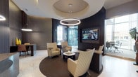 This single-floor space presents a thoughtfully designed interior that blends contemporary elegance with functional sophistication. The lobby area serves as the focal point upon entry, featuring striking marble-look flooring and a dramatic circular tray ceiling from which a large ring-shaped pendant light descends, casting a warm ambient glow over the seating area below. A curved dark wood accent wall anchors the space, housing a wall-mounted flat-screen display and providing a rich contrast to the lighter flooring and neutral-toned furnishings. The lounge seating area is arranged on a circular area rug, incorporating woven accent chairs and a low round cocktail table, creating an inviting, organized gathering spot. A dedicated reception desk and workstation area are visible beyond the lounge, finished with dark wood paneling and complementary pendant lighting. Adjacent to the main lobby, a formal conference room offers floor-to-ceiling glass walls that flood the interior with natural light and provide views of the surrounding streetscape. The conference room features a large polished stone-top table surrounded by executive chairs, accented by a striking starburst chandelier. Stone tile and warm wood wall treatments complete the refined, professional aesthetic throughout.