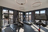 This fitness center in Salem, Massachusetts, offers a well-appointed, single-floor exercise space designed with both functionality and contemporary aesthetics in mind. The room is characterized by crisp white walls contrasted by a deep navy blue accent wall, creating a clean and energizing atmosphere suited for physical activity.

The space is equipped with a curated selection of cardio machines, including treadmills, an elliptical, and a stationary cycling bike with an integrated display screen. The flooring consists of dark rubberized interlocking tiles, providing durable, slip-resistant surfaces appropriate for high-impact exercise.

Overhead, a pair of circular ring pendant lights hang at varying heights, lending a modern, design-forward element to the space. The ceiling height contributes to an open, airy feel throughout the room. Large, floor-to-ceiling windows with dark frames line one wall, flooding the interior with ample natural light and offering views of the surrounding Salem streetscape and neighborhood.

Entry to the fitness center is through a full-glass-panel door with a dark metal frame, maintaining visual openness and continuity with adjacent spaces. The overall design balances practicality with a polished, modern sensibility, making it a functional and visually appealing environment for daily workouts.