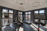 This fitness center in Salem, Massachusetts, offers a well-appointed, single-floor exercise space designed with both functionality and contemporary aesthetics in mind. The room is characterized by crisp white walls contrasted by a deep navy blue accent wall, creating a clean and energizing atmosphere suited for physical activity.

The space is equipped with a curated selection of cardio machines, including treadmills, an elliptical, and a stationary cycling bike with an integrated display screen. The flooring consists of dark rubberized interlocking tiles, providing durable, slip-resistant surfaces appropriate for high-impact exercise.

Overhead, a pair of circular ring pendant lights hang at varying heights, lending a modern, design-forward element to the space. The ceiling height contributes to an open, airy feel throughout the room. Large, floor-to-ceiling windows with dark frames line one wall, flooding the interior with ample natural light and offering views of the surrounding Salem streetscape and neighborhood.

Entry to the fitness center is through a full-glass-panel door with a dark metal frame, maintaining visual openness and continuity with adjacent spaces. The overall design balances practicality with a polished, modern sensibility, making it a functional and visually appealing environment for daily workouts.