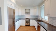 This single-floor space showcases a thoughtfully designed interior with a clean, contemporary aesthetic throughout. The kitchen is a standout feature, configured in a efficient U-shape layout with crisp white shaker cabinetry extending to the ceiling, dark countertops, and a light gray subway tile backsplash. Stainless steel appliances include a gas range with a wall-mounted range hood, a refrigerator, and a dishwasher. A window above the undermount sink fills the kitchen with natural light and provides views of the surrounding neighborhood.

The living area is generously proportioned and bathed in natural light through a large picture window that frames outdoor views. Warm hardwood-style flooring flows consistently throughout the living spaces and bedroom, complementing the bright white walls and clean crown molding details. The bedroom is a calm, neutral space with its own window and a decorative ceiling fixture.

The bathroom is finished to a high standard, featuring large-format hexagonal floor tiles in charcoal gray, white subway wall tiles with a decorative mosaic border, and a frameless glass-enclosed walk-in shower with dual shower fixtures. A window within the shower adds natural light to the space, reinforcing the unit's overall sense of brightness and openness.