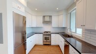 This single-floor space showcases a thoughtfully designed interior with a clean, contemporary aesthetic throughout. The kitchen is a standout feature, configured in a efficient U-shape layout with crisp white shaker cabinetry extending to the ceiling, dark countertops, and a light gray subway tile backsplash. Stainless steel appliances include a gas range with a wall-mounted range hood, a refrigerator, and a dishwasher. A window above the undermount sink fills the kitchen with natural light and provides views of the surrounding neighborhood.

The living area is generously proportioned and bathed in natural light through a large picture window that frames outdoor views. Warm hardwood-style flooring flows consistently throughout the living spaces and bedroom, complementing the bright white walls and clean crown molding details. The bedroom is a calm, neutral space with its own window and a decorative ceiling fixture.

The bathroom is finished to a high standard, featuring large-format hexagonal floor tiles in charcoal gray, white subway wall tiles with a decorative mosaic border, and a frameless glass-enclosed walk-in shower with dual shower fixtures. A window within the shower adds natural light to the space, reinforcing the unit's overall sense of brightness and openness.