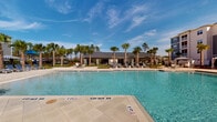 This pool facility in Panama City Beach, Florida, embodies a coastal resort aesthetic with thoughtfully designed outdoor and semi-enclosed spaces. The centerpiece is a generously sized swimming pool with crystal-clear turquoise water, marked with clearly designated shallow and deeper sections. The pool deck is finished in light-toned pavers and is lined with lounge chairs, framed by a backdrop of tall palm trees and open Florida sky.

The adjacent clubhouse structure is constructed with a distinctive light-toned natural stone masonry exterior and features a dark metal roof, lending a refined yet relaxed architectural character. Large aluminum-framed windows connect the interior to the outdoor spaces visually, filling the building with natural light while maintaining views of the pool and landscaping.

A covered outdoor terrace extends along the building's perimeter, furnished with bistro-style tables and navy blue and white woven chairs, creating a comfortable al fresco gathering space. Oversized white planters with lush tropical fan palms punctuate the terrace, enhancing the resort ambiance. The open-air design allows for unobstructed views of the surrounding greenery and manicured grounds, contributing to the overall breezy, vacation-inspired atmosphere of this Panama City Beach amenity space.