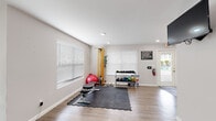 This fitness center is a thoughtfully organized, single-floor facility designed to support a full range of workout routines. The space features light wood-style flooring throughout, neutral-toned walls, and recessed lighting that creates a clean, energizing atmosphere. Large windows fitted with white horizontal blinds allow natural light to fill the room while maintaining privacy.

The facility is divided into two functional workout zones. The first area is dedicated to strength and functional training, featuring a rack of dumbbells and medicine balls, a weight bench, a stability ball, a resistance cable station, and interlocking foam floor mats that provide a safe surface for floor exercises. A wall-mounted flat-screen television is positioned for easy viewing during workouts, and posted fitness center rules ensure a structured environment.

The second zone is outfitted with cardio equipment, including a treadmill, an air resistance bike, and an elliptical machine, all arranged along a wall of windows that invite natural light. A large mirror spans one wall, enhancing the sense of openness and allowing users to monitor their form. The facility also includes safety equipment such as a fire extinguisher and clearly marked exit signage, reflecting a commitment to user safety and accessibility.