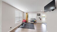 This fitness center is a thoughtfully organized, single-floor facility designed to support a full range of workout routines. The space features light wood-style flooring throughout, neutral-toned walls, and recessed lighting that creates a clean, energizing atmosphere. Large windows fitted with white horizontal blinds allow natural light to fill the room while maintaining privacy.

The facility is divided into two functional workout zones. The first area is dedicated to strength and functional training, featuring a rack of dumbbells and medicine balls, a weight bench, a stability ball, a resistance cable station, and interlocking foam floor mats that provide a safe surface for floor exercises. A wall-mounted flat-screen television is positioned for easy viewing during workouts, and posted fitness center rules ensure a structured environment.

The second zone is outfitted with cardio equipment, including a treadmill, an air resistance bike, and an elliptical machine, all arranged along a wall of windows that invite natural light. A large mirror spans one wall, enhancing the sense of openness and allowing users to monitor their form. The facility also includes safety equipment such as a fire extinguisher and clearly marked exit signage, reflecting a commitment to user safety and accessibility.