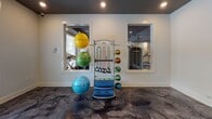 This single-floor fitness studio is thoughtfully designed to support a wide range of exercise activities. The space is accessed through elegant double French doors framed in white, accented by an overhead exit sign and set against crisp white walls — creating a clean, welcoming entry point.

The main exercise area features a stunning polished epoxy floor with a bold, marbled dark-grey aesthetic that immediately sets the tone for a high-end fitness environment. Large windows fitted with white horizontal blinds allow abundant natural light to flow in, offering views of lush outdoor greenery and a pool area beyond.

A dedicated spin cycling room is a standout feature, housing multiple red stationary bikes arranged in rows facing a wall-mounted flat-screen television set against a vibrant lime-green accent wall. Recessed ceiling lighting and a dark ceiling treatment give this room an energized, boutique studio feel.

The open fitness area is equipped with a centrally positioned equipment rack holding an assortment of medicine balls, resistance bands, and stability balls in multiple sizes and colors, adding a dynamic and functional element to the space. Interior pass-through windows connect the main exercise room to a separate cardio and strength equipment area, which is visible in the background and features treadmills and additional machines.

Throughout the studio, recessed lighting ensures consistent illumination, while the overall design balances functionality with modern aesthetic appeal.