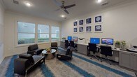 This single-floor business center is thoughtfully designed to accommodate a variety of work and meeting needs. The space is finished with crisp white walls accented by crown molding, and a bold grey and blue striped carpet that adds visual energy throughout. Recessed lighting and ceiling fans maintain a bright, comfortable atmosphere, while large windows along one wall invite natural light and provide views of the surrounding greenery.

The workstation area lines one wall with a row of desktop computers, including both PC and Mac setups, along with a shared printer—offering residents full connectivity for professional tasks. Adjacent to this zone, a comfortable lounge area features sleek black leather seating arranged around a metallic round coffee table, ideal for casual meetings or relaxed work sessions. A wall-mounted flat-screen TV and a water dispenser add convenience to the space.

A dedicated conference nook includes a round table with chairs, suited for small group discussions. A separate private room with glass-paneled doors provides an enclosed space for focused work or meetings. A glass whiteboard panel on the wall supports brainstorming and collaborative planning. A "Welcome Home" mat near the entry adds a welcoming touch to this versatile, professionally appointed space.