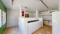Located at 10654 North 60th Avenue in Glendale, Arizona, this community laundry facility offers a clean, functional, and well-organized space designed to serve residents efficiently. The room is finished with durable, light-toned ceramic tile flooring throughout and features bright white walls, with one striking green accent wall that adds a bold visual element to the interior.

A row of commercial-grade top-loading washing machines is neatly arranged along a central white partition wall, providing ample laundry capacity for residents. Track lighting runs along the ceiling, ensuring the space is well-lit at all times. A wooden hanging rod is mounted along one wall, offering a convenient area for garment hanging, while a built-in countertop provides additional folding or sorting space.

The vivid green accent wall features several large square windows that frame views of the outdoor swimming pool and surrounding residential buildings, allowing natural light to filter into the space and creating a connection to the outdoor amenity areas. Decorative artwork, including a vibrant sunflower print, adds a welcoming touch to the otherwise utilitarian environment. A utility door and waste receptacle are also present, rounding out the practical amenities of this well-maintained shared laundry room.