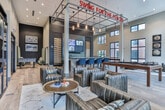 Located at 800 Battery Ave SE in Atlanta's vibrant Battery district, this single-floor amenity lounge is designed to impress with a sophisticated blend of industrial and contemporary aesthetics. Soaring ceilings and an abundance of oversized, black-framed windows flood the expansive open-plan space with natural light while offering views of the surrounding urban landscape.

Rich, wide-plank wood-look flooring anchors the entire space, complementing a carefully curated interior palette of deep navy, charcoal, and warm neutrals. A central bar and counter area features striking reclaimed wood paneling with a clean quartz surface, flanked by dramatic floor-to-ceiling wine display towers and mounted flat-screen televisions — ideal for social gatherings. Pendant lighting overhead adds an artisanal touch.

The lounge area is furnished with tufted leather club chairs and sofas arranged around a round wooden coffee table, accented by baseball-themed wall art that nods to the space's proximity to Truist Park. A billiards table provides a focal point for recreational use, while glass-paneled double doors connect seamlessly to an outdoor terrace, extending the entertaining space. Discrete enclosed rooms along the perimeter offer additional flexibility. The overall ambiance is warm, energetic, and thoughtfully designed for both relaxation and socializing.