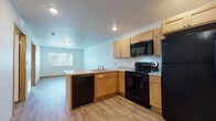 This single-floor unit offers a practical, well-equipped layout designed for comfortable everyday living. The open-concept design connects the kitchen and living area seamlessly, creating a spacious, airy feel enhanced by recessed lighting and neutral light-blue and white walls throughout.

The kitchen is a functional centerpiece, featuring light wood cabinetry with ample upper and lower storage, a full suite of black appliances including a refrigerator, electric range, over-the-range microwave, and dishwasher. An L-shaped countertop layout with a built-in sink anchors the space, while a peninsula counter with additional workspace partially separates the kitchen from the living area. Light-toned vinyl plank flooring extends throughout the main living spaces, adding warmth and easy maintenance.

The bedroom is generously sized, fitted with a ceiling fan, a window allowing natural light, and direct access to adjacent rooms. The bathroom features a clean, all-white walk-in shower unit with a curtain rod and a chrome towel bar, complemented by wood-toned baseboards and matching vinyl flooring.

A dedicated in-unit laundry area houses a stacked washer and dryer, providing added convenience. The entry door features a keypad lock, offering a modern security feature. Overall, the space presents a clean, move-in ready environment with practical finishes throughout.