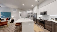 Located at 2332 N 7th St in Phoenix, AZ, this well-appointed two-bedroom, two-bathroom residence offers a thoughtfully designed single-level layout with modern finishes throughout. The open-concept living, dining, and kitchen area serves as the heart of the home, anchored by a substantial island with a white quartz countertop, an undermount sink, and bar seating. The kitchen is outfitted with sleek white upper cabinetry, warm wood-toned lower cabinets, a white subway tile backsplash, and a full suite of stainless steel appliances. Light wood-style flooring flows seamlessly throughout the space, complementing the neutral gray walls and recessed lighting overhead. The living area offers a comfortable retreat, highlighted by large windows with draping curtains that allow natural light to filter in. The primary bedroom is a serene space with room for a king-sized bed, while the second bedroom features a bright window and ample natural light. Both bathrooms are finished with modern vanities featuring dark wood cabinetry and white countertops. The primary bath includes a glass-enclosed walk-in shower with white tile surround, while the secondary bath offers a full tub-shower combo. Closets and hallways provide practical storage throughout this well-balanced residence.