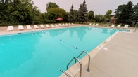 This recreational facility centers around a generously sized outdoor swimming pool, featuring vivid turquoise water and clearly marked depth indicators along its walls. The pool is equipped with stainless steel entry handrails and a tiled entry step area, making access straightforward and safe. A lane divider runs across the pool, accommodating both recreational and lap swimming. The surrounding concrete pool deck is well-appointed with numerous white lounge chairs arranged along the perimeter, along with colorful shade umbrellas providing relief from the sun. A perimeter fence encloses the pool area, ensuring safety and privacy, while mature trees—including tall evergreens—frame the backdrop, creating a natural, park-like atmosphere. A distinctive Tudor-style clubhouse with a steeply pitched roof, dark wood trim detailing, and brick exterior anchors one end of the property, providing functional support facilities. Complementing the outdoor amenities, an indoor racquetball court features a polished hardwood floor with regulation court markings, bright white walls, and overhead skylight panels that provide clean, even illumination throughout the space.