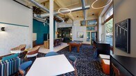 Located at 221 Fifth Street East in downtown Saint Paul, this single-floor amenity lounge delivers a polished, contemporary atmosphere that blends urban energy with comfortable gathering spaces. The interior is characterized by soaring ceilings with exposed ductwork and structural columns, lending an industrial-chic aesthetic softened by warm wood-framed windows and circular pendant lighting.

Large, gold-trimmed windows line the perimeter of the space, flooding the interior with abundant natural light and framing views of the surrounding historic downtown streetscape, including classic brick architecture and tree-lined sidewalks. The open layout accommodates multiple distinct zones, including a lounge area anchored by a navy sectional sofa, colorful accent pillows, and a bold patterned carpet in deep navy and white tones.

A full-size billiards table serves as a central focal point, complemented by high-top bar seating and a wet bar with warm wood cabinetry. A media lounge area features a wall-mounted flat-screen television set against textured tile, flanked by directional sconce lighting and an orange upholstered ottoman. White communal tables with modern chairs round out the space, making it well-suited for casual socializing, recreation, and community gatherings.