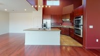 This single-floor residence makes a strong design statement through its consistent use of deep red accent walls, rich cherry-toned hardwood flooring, and dark wood cabinetry throughout. The open-concept kitchen is a standout feature, boasting soaring double-height ceilings with clerestory windows that flood the space with natural light. A large central island with a granite countertop and double sink serves as the kitchen's focal point, complemented by stainless steel appliances including a French door refrigerator, gas cooktop, double wall ovens, and rich wood cabinetry. The living area extends from the kitchen with gleaming hardwood floors and sliding glass doors that open to an outdoor view, inviting abundant natural light. Bedrooms are carpeted and well-proportioned, with one featuring striking red accent walls and soaring ceilings, while another offers neutral tones and a bright window. A spacious walk-in closet with wrap-around shelving provides generous storage. The bathroom is appointed with large-format terra cotta tiles, a soaking tub with full tile surround, and dark wood vanity cabinetry. A dedicated laundry area with side-by-side front-loading washer and dryer and overhead cabinetry completes this well-appointed residence.
