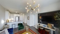 This single-floor space in The Colony showcases a thoughtfully designed open-concept layout that seamlessly connects the kitchen and living areas. The kitchen features white shaker-style cabinetry, stainless steel appliances, and a peninsula with pendant lighting, offering both functionality and visual appeal. Dark hardwood-style flooring runs throughout, grounding the space with warmth and contrast against the crisp white walls and ceilings.

The living area is anchored by a striking sputnik-style chandelier with exposed Edison bulbs, serving as a bold focal point that elevates the room's character. A large chalkboard-style accent wall frames the media area, adding a creative, personalized touch to the space. Eclectic furnishings, including a rich green velvet accent chair, a patterned area rug, and woven storage baskets, contribute a layered, curated aesthetic.

The layout flows openly toward interior doors, suggesting additional private spaces beyond. Natural light filters in, brightening the neutral palette. Overall, the space balances modern design sensibilities with distinctive, playful details that give it a unique and inviting atmosphere.
