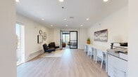 Located at 7288 Maple Street in Mission, BC, this single-floor amenity space offers a clean, contemporary design that blends functionality with modern aesthetics. The layout is divided into two distinct areas — a living room and a dining room — unified by light-toned hardwood-style flooring, recessed lighting, and a crisp white palette throughout.

The living room features a welcoming seating arrangement with sleek black armchairs set on a area rug, complemented by decorative wall clocks and curated artwork. A long communal counter with white chairs lines one wall, offering an additional workspace or casual gathering spot. A black-framed glass partition door leads into a sunlit lounge area furnished with plush, rounded chairs set against expansive floor-to-ceiling windows that flood the space with natural light and frame views of the outdoor surroundings.

The dining area is anchored by a striking marble-topped table surrounded by white chairs, with a large window allowing natural light to pour in. A built-in kitchenette with light wood cabinetry and a sink provides convenient utility. Bold abstract artwork adorns the walls throughout, adding visual interest and a sophisticated ambiance. The overall atmosphere is bright, airy, and thoughtfully designed for both comfort and versatility.