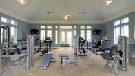 This fitness center offers a thoughtfully designed workout environment across a single floor, combining functionality with a bright, open atmosphere. The space is painted in a calming slate blue, which pairs elegantly with the crisp white trim and neutral carpeted flooring throughout. Large windows and transom windows line the walls, flooding the room with natural light and offering views of the surrounding greenery, while a set of French doors at the rear provides both visual openness and easy access to the outdoors. A ceiling fan with a light fixture overhead ensures comfortable air circulation during workouts. The equipment layout is well-organized, featuring a range of cardio machines including ellipticals, stationary bikes, and treadmills, alongside strength and resistance training equipment such as a weight bench and cable machine. Two wall-mounted flat-screen televisions are positioned on opposite sides of the room, providing entertainment options for users during their sessions. The overall ambiance is clean, airy, and inviting, making this fitness center a practical and pleasant space for a comprehensive workout routine.