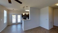 This single-floor space is designed with an open-concept layout that seamlessly connects the living area to the kitchen. The flooring throughout is a warm hardwood-style material that adds continuity and visual cohesion across the space. The living area is equipped with a ceiling fan and benefits from natural light that flows in from the French doors, which open to a private outdoor balcony with railing, offering a pleasant outdoor connection.

The kitchen is defined by a peninsula with a light-colored countertop that serves as both a functional workspace and a natural divider between the kitchen and living areas. Dark espresso-toned cabinetry provides a strong visual contrast against the white walls and light countertop. Stainless steel appliances, including a microwave and range, are built into the cabinetry for a streamlined look. Three pendant lights with warm-toned shades hang above, adding focused task lighting and a decorative touch. The overall ambiance is clean, modern, and bright, with neutral tones and practical finishes throughout.