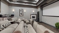 This private home theater is thoughtfully designed to deliver a true cinema experience within a refined, contemporary setting. The room is finished in a sophisticated neutral palette of gray and taupe, with soft carpeting underfoot and a coffered ceiling detail that adds architectural interest while housing the overhead projector and ventilation systems.

Seating is arranged in tiered rows of plush, oversized suede-tone sofas, each accented with geometric patterned throw pillows. Built-in armrest lighting provides subtle illumination without disrupting the viewing atmosphere. The rows are elevated stepwise from front to back, ensuring clear sightlines to the large wall-mounted projection screen at the front of the room.

A raised bar area along the rear wall offers a more casual viewing option, furnished with sleek chrome and black leather swivel stools arranged around high-top tables. This area is further distinguished by a gallery wall of framed black-and-white Hollywood portrait photography, lending the space a classic, cinematic glamour.

Wall sconces throughout provide warm ambient lighting, and in-wall speaker panels suggest a full surround sound audio system. A small potted plant near the entrance adds a touch of organic warmth to the otherwise polished, entertainment-focused environment.